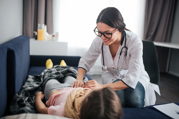 Positives und fröhliches Lächeln der jungen Ärztin. Sie schaut nach unten und berührt den Bauch des Mädchens. Doktor überprüfen Sie es. Kind liegt gerade auf dem Bett.