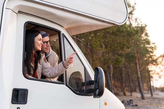 Positives verliebtes Paar, das mit dem Finger zeigt, während es während eines Picknicks im Wald am Auto sitzt und vor der Kamera lächelt