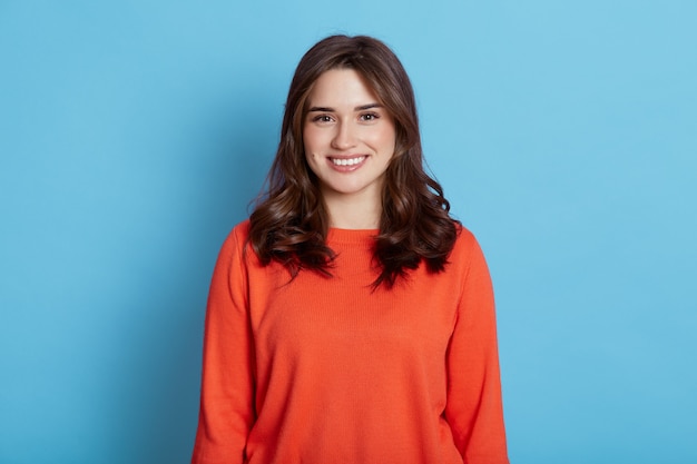 Positivo alegre joven hermosa mujer con cabello oscuro vistiendo ropa casual, mirando y sonriendo agradablemente, vistiendo un suéter naranja, de pie aislado sobre una pared azul.