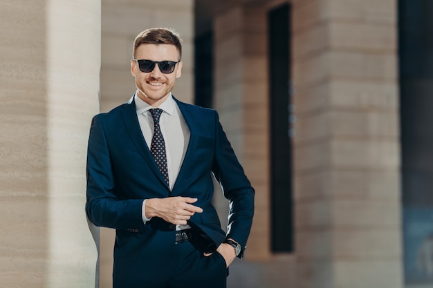 Positivo, alegre, joven, macho, usa, traje formal, usa tonos modernos, mantiene la mano en el bolsillo