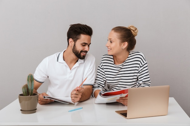 positivo alegre joven pareja amorosa amigos hombre y mujer sentados en la mesa en el interior aislado sobre la pared gris usando la computadora portátil leyendo un libro estudiando.