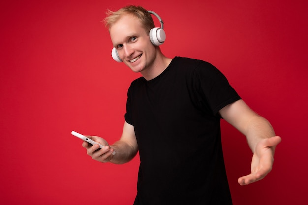 Positivo encantador guapo rubio joven vestido con camiseta negra y auriculares blancos de pie