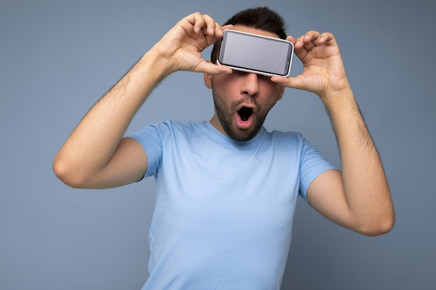 Foto positivo espantado jovem bonito com barba por fazer com barba vestindo camiseta azul todos os dias, isolado sobre fundo azul, segurando e mostrando o telefone móvel com visor vazio para maquete.