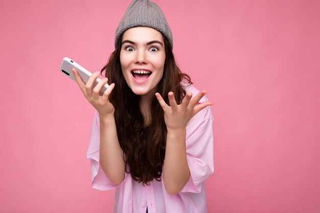 Positivo feliz atractiva joven morena mujer vistiendo elegante camisa rosa y sombrero gris aislado sobre