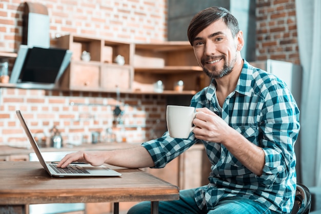 Positivo, feliz, bom, sentado na cozinha e sorrindo enquanto saboreia seu chá