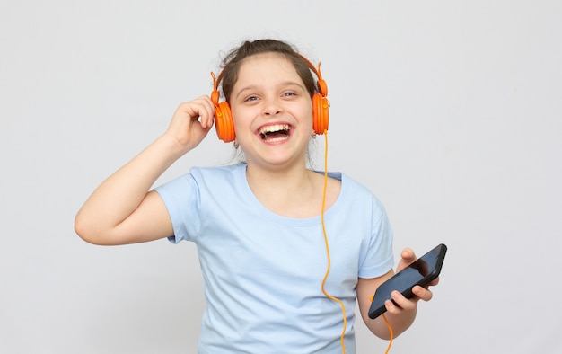 Positivo hermosa niña caucásica vistiendo jeans en general sobre fondo blanco tiene teléfono celular moderno conectado a auriculares, aprieta el puño de buenas emociones, exclama con alegría,