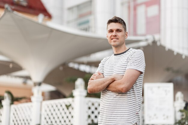 Positivo jovem bonito com um lindo sorriso em uma elegante camiseta listrada está de pé na rua da cidade.