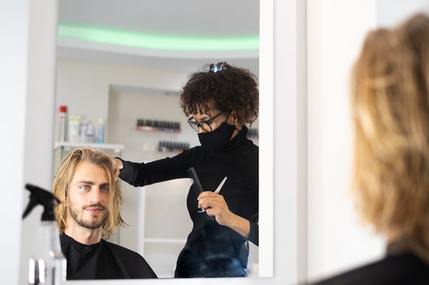 Positivo jovem cabeleireiro negro gostando de trabalhar no salão de beleza e secar o cabelo da cliente