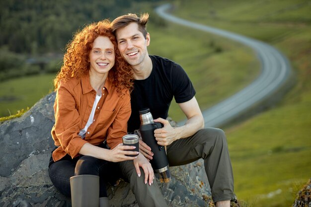 Positivo jovem casal caucasiano senta-se fazendo uma pausa para descansar nas montanhas, bebendo chá da garrafa térmica.