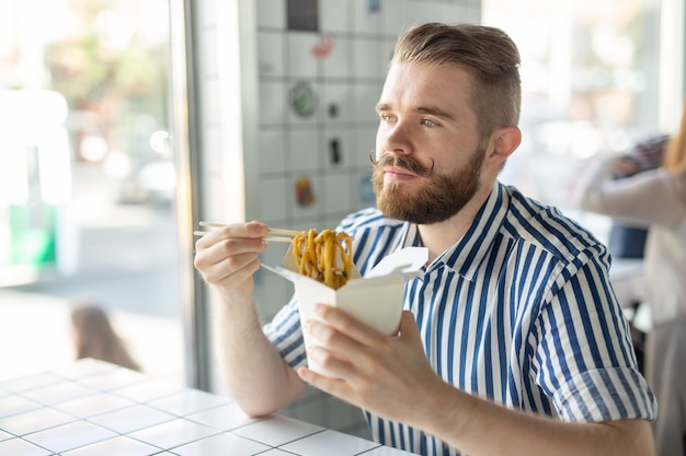 Positivo jovem elegante comendo macarrão chinês em um café durante uma pausa no trabalho. O conceito de descanso e lanche saudável.