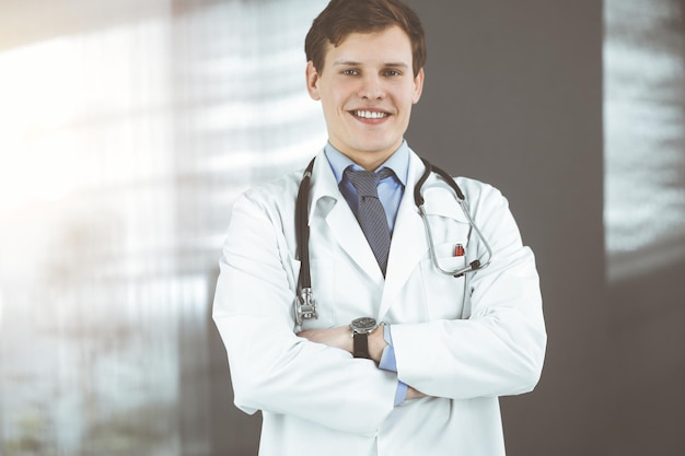 Positivo jovem médico está sorrindo para a câmera. Retrato de médico profissional na clínica ensolarada.