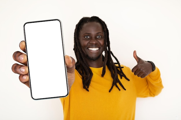 Positivo jovem negro segurando smartphone em branco e mostrando o polegar para cima gesto