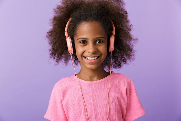 positivo otimista feliz jovem garota africana posando isolado sobre a parede roxa, ouvindo música com fones de ouvido.