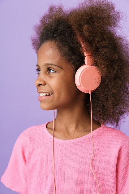 positivo otimista feliz jovem garota africana posando isolado sobre a parede roxa, ouvindo música com fones de ouvido.