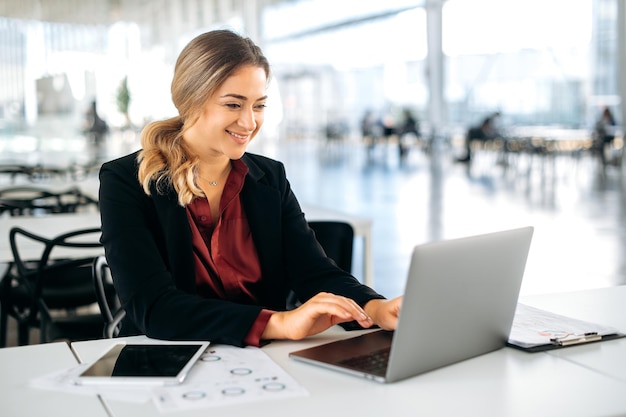 Foto positivo satisfeito bem sucedido influente mulher de negócios caucasiana gerente de produto em roupas elegantes diretor da empresa senta-se em uma mesa de trabalho no escritório trabalhando em um laptop sorri