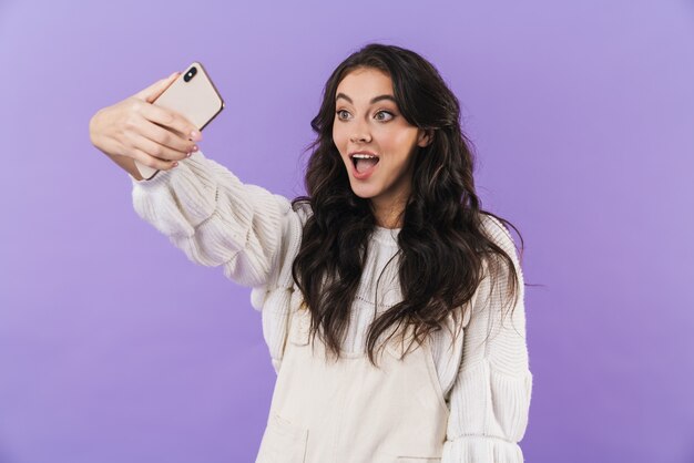 positivo sorprendido linda joven morena posando aislada sobre la pared de la pared púrpura tomar un selfie por teléfono móvil.