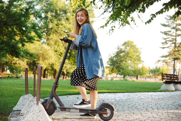 positivo sorprendido optimista feliz adolescente en el parque caminando en scooter con teléfono móvil.