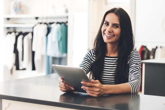 Foto posso ajudar? mulher jovem e bonita segurando um tablet digital e olhando para a câmera com um sorriso enquanto está na loja de roupas