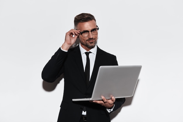 Posso te ajudar? Jovem bonito com terno completo usando laptop e olhando para a câmera com um sorriso em pé contra um fundo branco