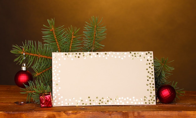 Postal en blanco, bolas de Navidad y abeto en mesa de madera sobre fondo marrón