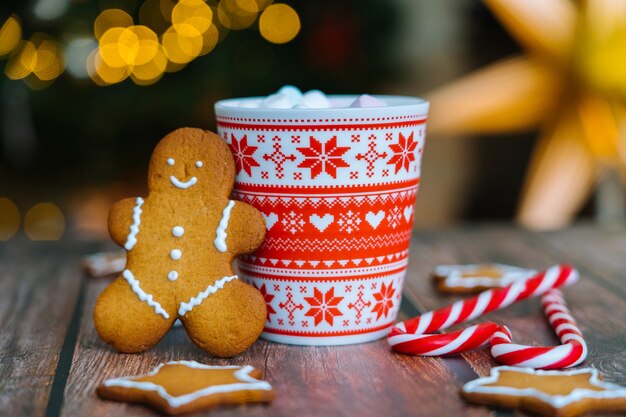 Postal de Natal com caneca vermelha do homem-biscoito com enfeites de natal na luz bokeh da árvore de Natal e doces listrados vermelhos de férias.