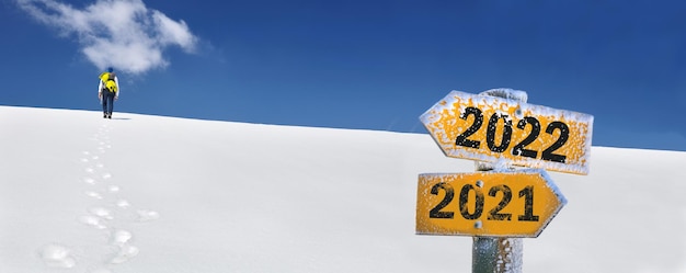 Posten Sie das Schild Richtung 2022 in Richtung eines Mannes, der schneebedeckte Berge erklimmt und 2021 gegenüber