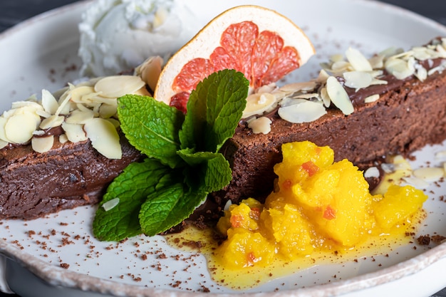 Postre de chocolate brownie con una bola de helado blanco, rebanadas de mango y una rodaja de pomelo. En el fondo hay un capuchino. Sobre un fondo oscuro