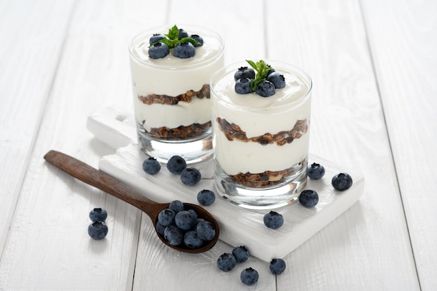 Postre con requesón arándanos frescos y granola en un vaso sobre un fondo de madera blanca