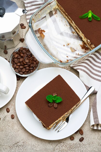 Postre tiramisú italiano tradicional en una fuente para hornear de vidrio y la porción en la mesa de hormigón gris