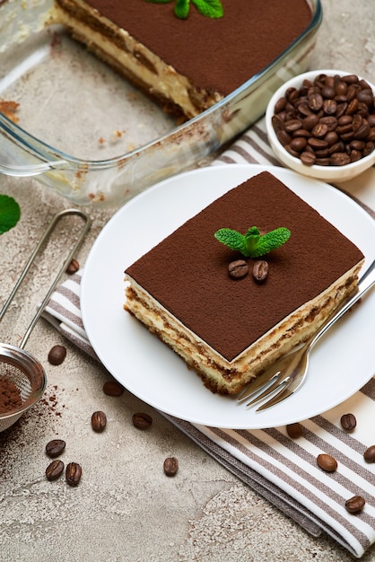 Postre tiramisú italiano tradicional en una fuente para hornear de vidrio y la porción en la mesa de hormigón gris