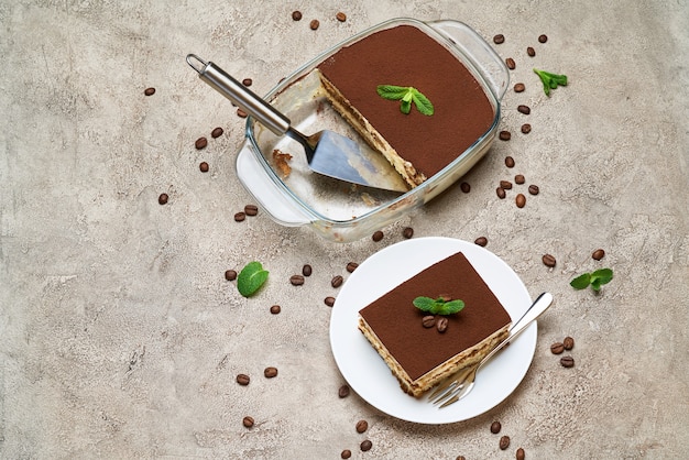 Postre tiramisú italiano tradicional en una fuente para hornear de vidrio y la porción en la mesa de hormigón gris