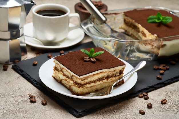 Postre tiramisú italiano tradicional en una fuente para hornear de vidrio y la porción en la mesa de hormigón gris