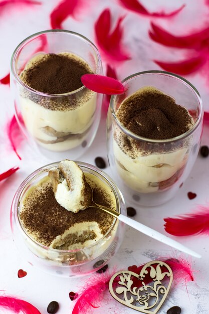 Postre tiramisú en vasos de doble fondo sobre una superficie blanca entre plumas rojas