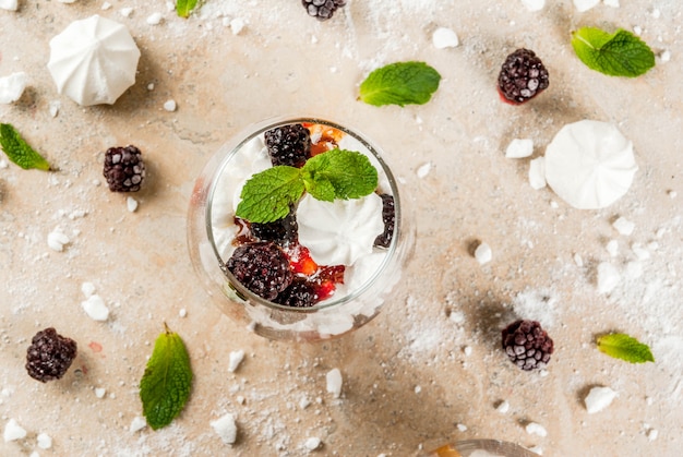 Postre tradicional inglés. Eton mess: crema batida, merengue, moras frescas, salsa y caramelo. En servir vasos en una mesa de piedra clara. Copia espacio vista superior