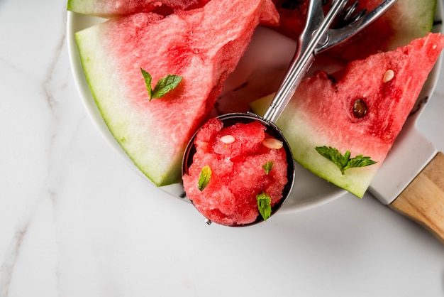 Postres de frutas de verano, cócteles congelados. Helado de granito de sandía con menta, una cucharada en una cuchara para helado, en un plato con rodajas de sandía.