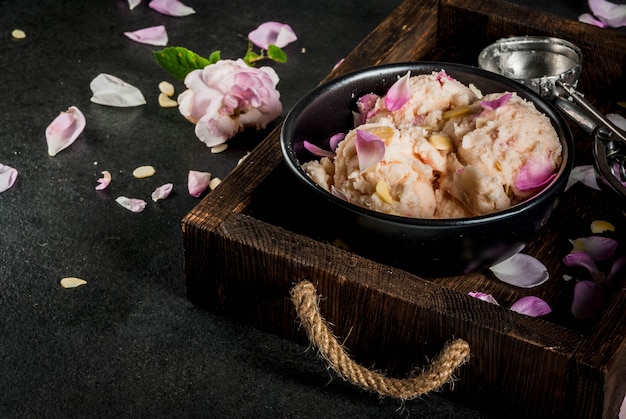 Postres refrescantes de verano. Comida vegana de dieta. Helado con pétalos de rosa y rodajas de almendras, en bandeja de madera vieja, en la mesa de piedra negra. Con cuchara para helado e ingredientes.