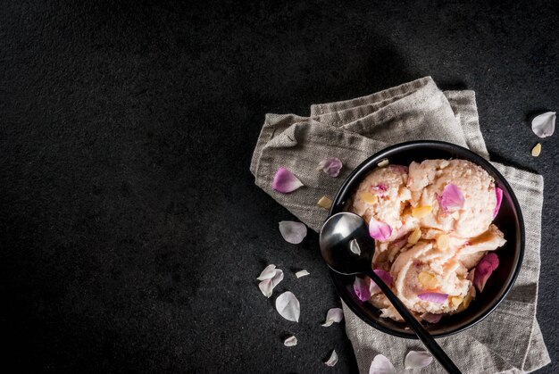 Postres refrescantes de verano. Comida vegana de dieta. Helado con pétalos de rosa y rodajas de almendras, en bandeja de madera vieja, en la mesa de piedra negra. Con una cuchara en un tazón negro. Copia espacio vista superior