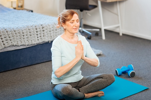 Postura de lótus. Mulher simpática de cabelos claros e cabelos presos meditando na postura de lótus enquanto está sentada no tapete azul