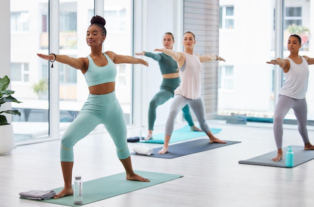 La postura del guerrero del yoga y las mujeres con el entrenador en la clase para el ejercicio, la aptitud y el equilibrio Las chicas de yoga y la práctica en grupo para el bienestar holístico, el cuerpo saludable y el estiramiento muscular para la flexibilidad en el gimnasio