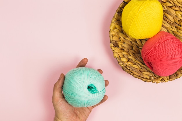 Postura plana da mão de uma mulher segurando uma linda bola de menta verde de algodão ao lado de bolas de rosa cor de rosa e amarelas escuras de algodão em uma cesta com fundo rosa pastel e espaço de cópia