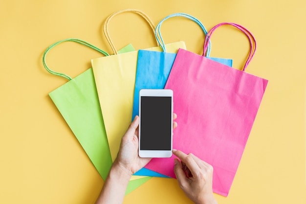 Postura plana das mãos da mulher usando o smartphone com bolsas coloridas em fundo amarelo