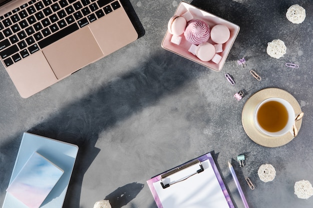 Postura plana de artigos de papelaria de escritório com uma xícara de chá com marshmallow e laptop em um fundo cinza com sombras.