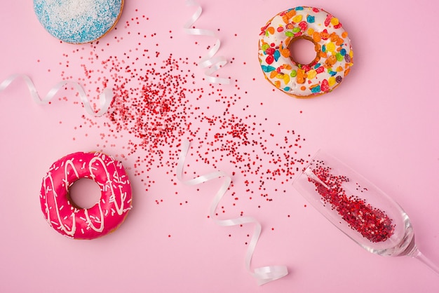 Foto postura plana de celebração. taça de champanhe com flâmulas de festa coloridas e deliciosos donuts no fundo rosa.