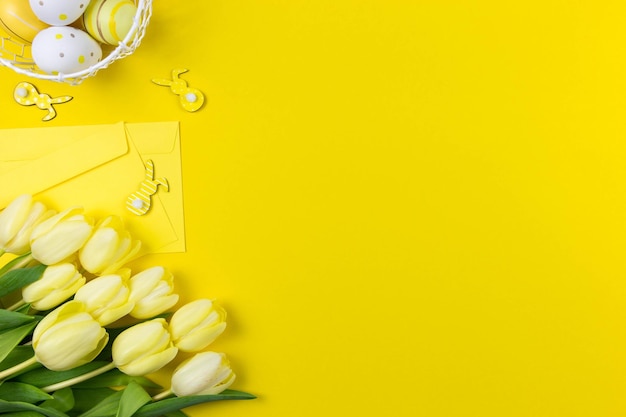 Postura plana de páscoa com tulipas amarelas ovos de páscoa coloridos decoração de coelhinhos e envelopes em fundo amarelo Vista superior Copiar espaço