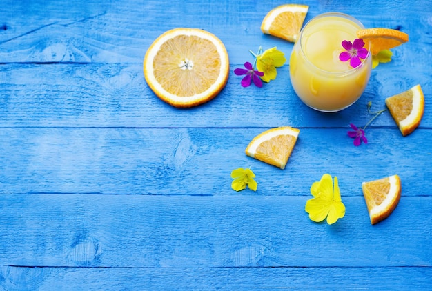 Postura plana de suco de laranja fresco guarnecido com uma fatia de laranja madura e flores na mesa de madeira azul