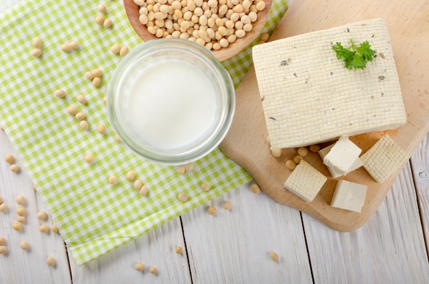 Postura plana em alternativas Nondairy Leite de soja ou iogurte em frasco de pedreiro e tofu na mesa de madeira branca com soja em tigela de lado