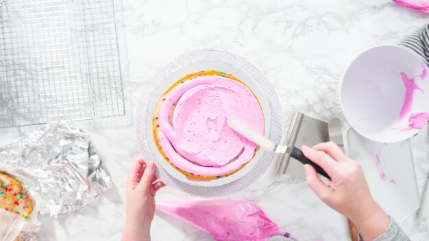 Postura plana. Passo a passo. Cobertura de camadas de bolo funfetti redondo com cobertura de creme de manteiga rosa.