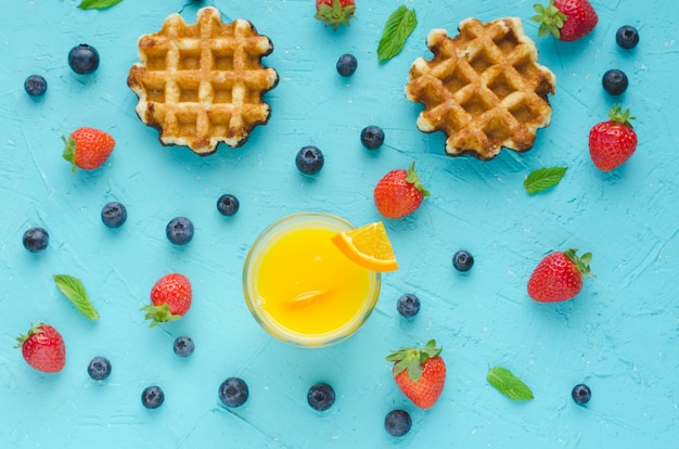 Postura plana. waffles, suco de laranja, frutas vermelhas e folhas de hortelã na superfície azul