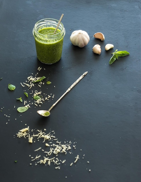 Pote de pesto caseiro fresco com ingredientes para ele folhas de manjericão fresco queijo parmesão ralado e alho sobre pano de fundo preto Vista superior