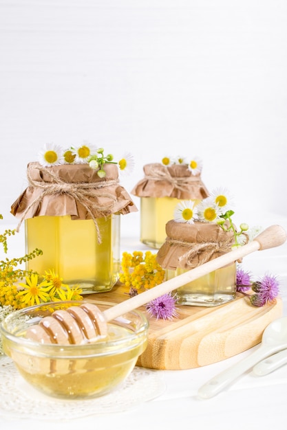 Potes de miel líquida fresca completa y palo de miel con flores silvestres de verano en mesa blanca